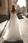 A woman viewed from behind wearing a long-sleeve white wedding gown with a lace bodice and a full tulle skirt, standing on a sunlit city street with buildings and parked cars in the background.