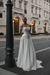 A woman modeling an off-the-shoulder white wedding gown with a fitted corset-style bodice and a flowing skirt, standing on a city street beside a black lamppost with a grand building in the background.