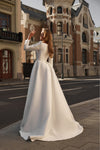 A woman wearing a long-sleeved white wedding gown stands on a city street corner with historic brick buildings in the background.