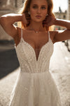 A woman wearing a white beaded wedding dress with thin straps and a deep V-neckline, standing outdoors in warm sunlight.