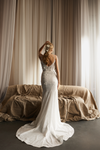 A woman stands with her back to the camera wearing a fitted white wedding gown with a long train, posed in front of beige curtains and a draped sofa in a softly lit indoor setting.