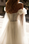 A woman wearing a white off-the-shoulder tulle dress, shown from the back in warm sunlight. The dress features a sheer corset-style back with a chain detail and large ruffled fabric flower details on the shoulders.
