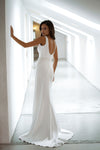 A woman wearing a sleeveless white wedding gown with a low open back and fitted silhouette, standing in a bright white corridor and looking back over her shoulder.