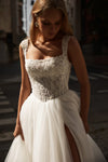 A woman wearing a white sleeveless wedding dress with a heavily beaded, textured bodice and a flowing tulle skirt with a thigh-high slit, photographed outdoors on a sunlit street.