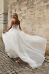 A woman wearing a strapless white wedding gown with a sheer lace bodice and a voluminous flowing skirt, standing on a cobblestone street beside a stone wall.