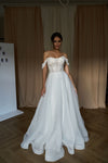 A woman modeling an off-the-shoulder white wedding gown with a structured lace bodice and a full flowing skirt, standing in front of beige curtains and a wood floor.