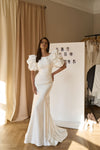 A woman models a floor-length white wedding gown with dramatic puffed short sleeves and a fitted silhouette, standing indoors in a room with beige curtains, a white photo board, and a clothing rack in the background.
